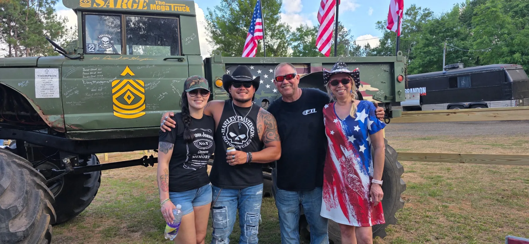 Four people smiling in front of Sarge, a green military-style mega truck covered in veterans' signatures, with American flags behind